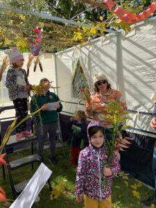 2 adults and 2 children hold fall themed decorations (flowers, straw, etc.). An open roof covered with loose green and yellow foliage is visibe at the tip of the image. Two white canvas walls lead down to green grass. The individuals are dressed for cool weather and are facing the camera.